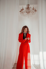 teen fashion. young blonde girl in modern red suit is standing casual, fastening the sleeve and looking down with smile on a white wall background with crystal chandelier. fashion concept, free space