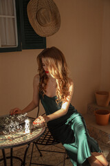 young blonde teen girl in green silk dress sits elegance with hands on table and looks down with smile on the italian patio background with window and straw hat. travel lifestyle concept, free space