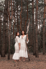 two brunette girls in white boho dresses are standing close with high wooden staff in hand and looking straight on pine forest background. female witch cosplay. lifestyle shaman concept, free space