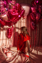 happy birthday girl. young glamour woman in red fur coat sits on floor with smile near rose cake on pink cloth background with heart balloons. disco valentine's party. celebration concept, free space