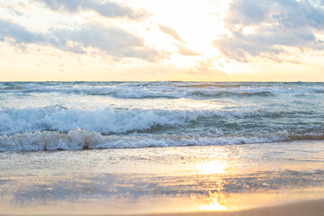 Fototapeta premium Golden sunlight shining through clouds over calm ocean with soft waves lapping at sandy beach