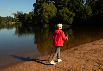 Grey-haired elderly woman walking with tracking sticks on the beach near lake.
