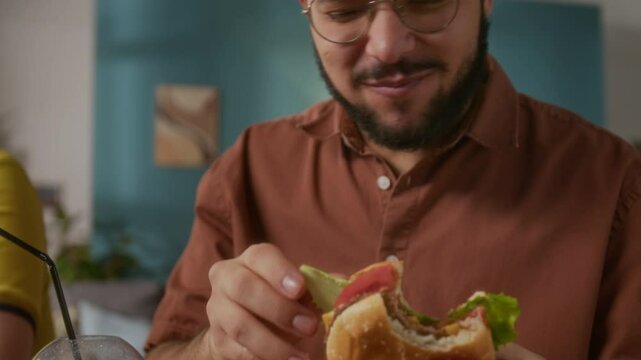 Tilt-down shot of young bearded man in glasses enjoying delicious burger and orange juice at home in living room