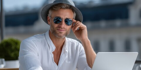 Man in a white shirt and sunglasses, adjusting his hat while using a laptop outdoors.