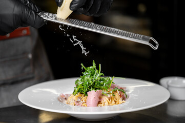 A chef grates cheese over a plate of pasta with arugula and ham, adding the finishing touch to a gourmet dish. The close-up captures culinary craftsmanship and fresh ingredients.