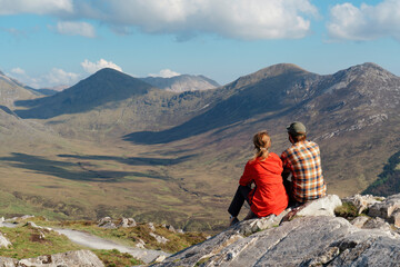 A couple sits on a rocky outcrop of Diamond Hill in Connemara National Park, admiring the sweeping...
