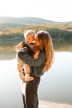 Happy woman holding baby girl 1 - 2 year old wearing knitted clothes having fun over lake and nature background. Motherhood. Autumn season.