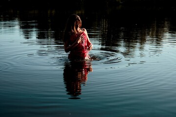 Beautiful brunette in a red dress stands waist-deep in a dark lake, gently splashing water on...
