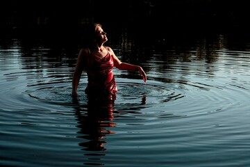Fototapeta premium A young woman in a red dress stands in a lake, her face turned upwards to the sky, with her right hand gently touching the surface of the water, creating a serene and contemplative mood.