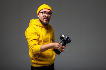 Smiling young man in yellow hoodie and beanie holding a camera in studio. Hipster photographer with beard, glasses, and tattoo on gray background.