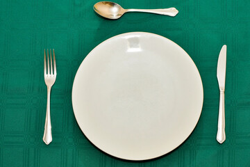 Close up view of a table setting with large white plate, knife, fork and spoon on a dark green tablecloth.