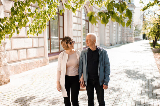 Senior happy couple of stylish man and woman 60-65 year old walking together on streets at city outdoors. Wellness lifestyle. Looking at each other.