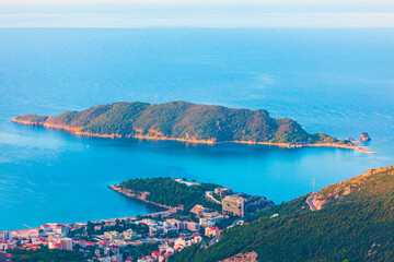 Scenic view of Becici on Budva Riviera, with a coastal town nestled at the base of green hills, overlooking calm blue waters of Adriatic Sea. In distance the lush Sveti Nikola Island rises from sea