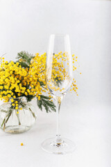 A close-up of a champagne flute reflecting yellow mimosa flowers from a glass vase, creating a delicate and artistic floral composition.