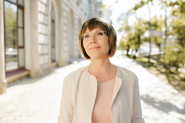 Smiling blonde senior woman 55-60 year old wearing eye glasses over urban city background close up. Mature lifestyle.