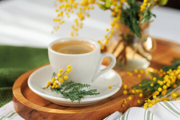A cozy spring morning scene with a white coffee cup on a wooden tray, decorated with fresh mimosa flowers, exuding warmth and charm.