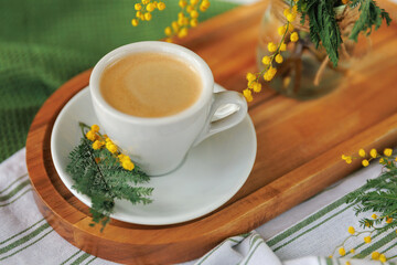 A cup of freshly brewed coffee on a wooden tray, decorated with yellow mimosa flowers, creating a bright and cozy spring mood.