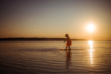 Fototapeta premium Silhouette of a girl running on water against the background of an evening sunset. Water splashes in all directions. Carefree baby fun. Happy healthy childhood.