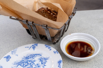 A small wire basket with brown paper is filled with pumpernickel bread. Next to the food is a small round white dish filled with balsamic vinegar dipping oil. A white and blue side plate is empty. 