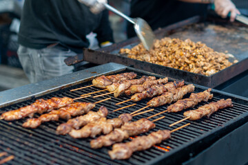 A man wearing a black t-shirt and jeans uses a barbecue utensil to grill boneless chicken. The charcoal barbeque has a bright flame. There's Korean chicken and spices on the wooden skewers grilling. 