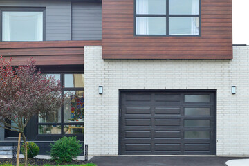 A modern black metal exterior garage door has four small horizontal glass windows. The luxury dark grey contemporary house has faux horizontal wood siding, white brick, grey walls, and glass windows. 