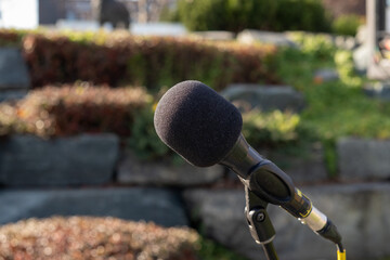 A black metal microphone on a stand. The round sponge ball style mic is a classic for concerts and evening music. The electronic equipment has a yellow cord. The background is an outdoor park. 