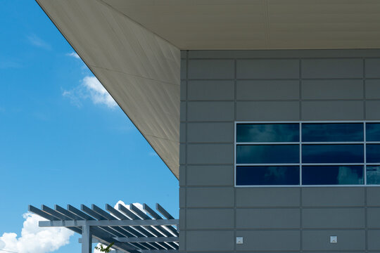 The corner eave of a grey aluminium composite panel waterproof modern building with a white eave and overhang. The exterior clad wall has multiple blue glass windows reflecting blue sky and clouds.