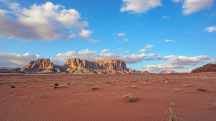 Wadi Rum desert with red sand lit by evening sun, rocky mountain formations in background