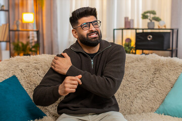 Indian man at home holds shoulder with tension, showing pain on his face while sitting on sofa....
