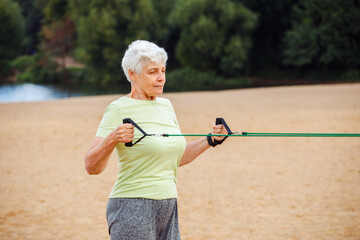 Old woman wear sportswear doing exercises outdoor on the beach in the morning using resistance...