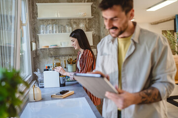 man stand in the kitchen and use tablet while woman washes the dishes