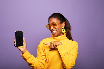 Portrait of positive afro american woman wearing yellow sweater and sumglasses promoter show smartphone over purple background.