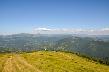 Fototapeta premium Rural house at the Beautiful mountain meadow beneath a sunny sky, surrounded by green hills and valleys, evoking tranquility. Carpathian Mountains, Ukraine