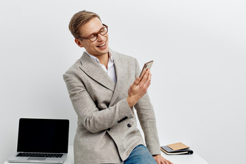 smiling man with smartphone wearing gray jacket casual pants sitting near laptop digital nomad...