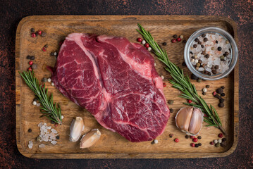Raw Rib Eye steak with spices on a wooden board on a dark background. View from above.