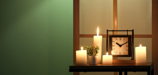 Burning candles and clock on table near green wall in dark room. Banner for design