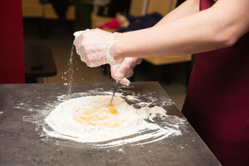 Making fresh pasta dough with eggs and flour. Gloved hands mix eggs and flour on a countertop,...