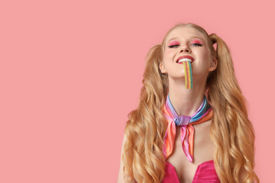 Beautiful young woman dressed as doll eating jelly candy on pink background