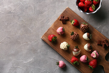 Wooden board with chocolate covered strawberries and bowl of fresh berries on grey background