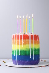 Delicious rainbow birthday cake with burning candles and sprinkles on light grey background