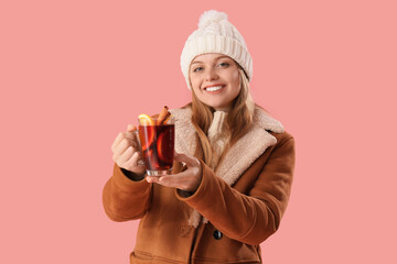 Beautiful young woman in warm clothes with glass cup of hot mulled wine on pink background