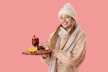 Beautiful young woman in warm clothes holding tray with glass cup of hot mulled wine on pink...