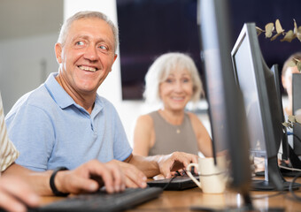 Positive interested elderly man using computer to surf internet in library. Concept of integration of older generations with modern technology..