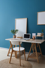 Workplace with laptop and blank posters near blue wall in room