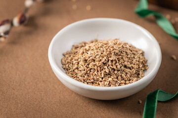 Fennel seeds in a white bowl on a textured brown surface with a delicate feather and blurred plant stem in the background