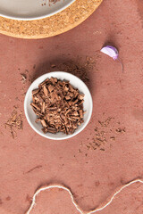 An overhead view features a bowl of chocolate shavings, scattered pieces, purple flower petals, and twine on a rustic reddish-brown surface.