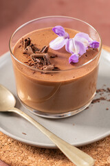 An inviting close-up of chocolate mousse in a glass, perfectly garnished with chocolate shavings and purple flowers on a plate with a spoon.