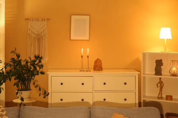 Chest of drawers with candles and glowing lamps in interior of living room