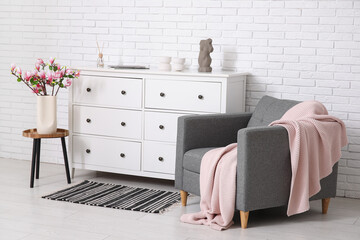 Chest of drawers, armchair and coffee table with blooming magnolia branches in vase near white brick wall in room © Pixel-Shot