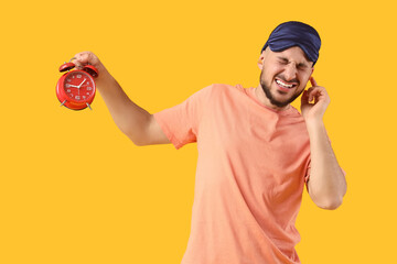 Young man with sleeping mask and alarm clock suffering from loud noise on yellow background